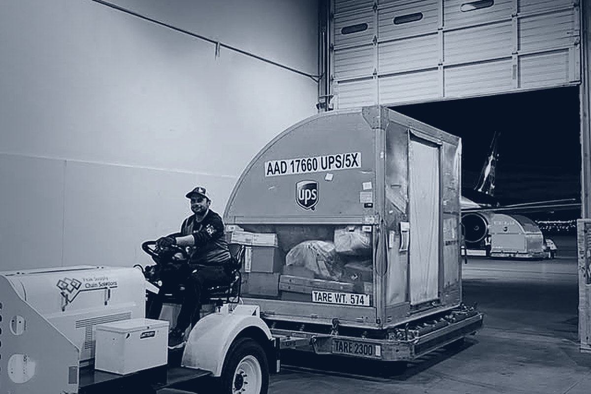 A Peak crew member driving a tug pulling a loaded ULD inside a Peak dock, with an aircraft visible beyond