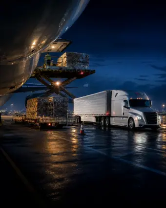 A branded tractor-trailer staged beside an aircraft cargo loader on a wet pre-dawn ramp — portrait framing for phones